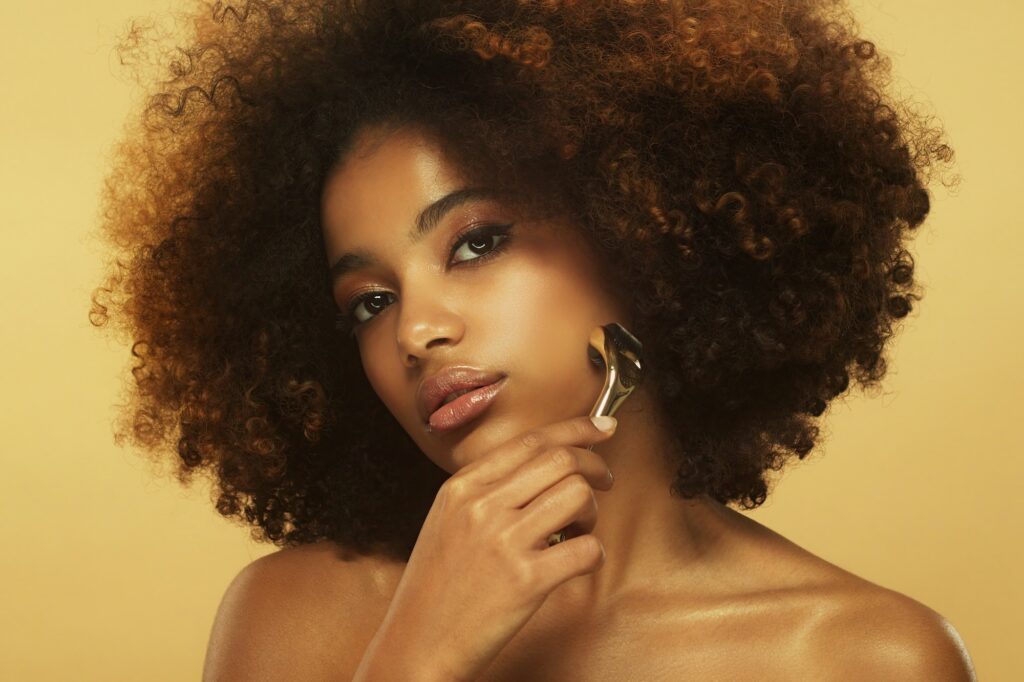 Young beautiful african woman with afro hairstyle using face roller for facial massage on her cheek